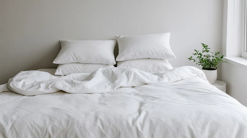 White bed with crumpled linen bedding and stacked pillows in a minimalist bedroom near a window with a potted plant on a side table