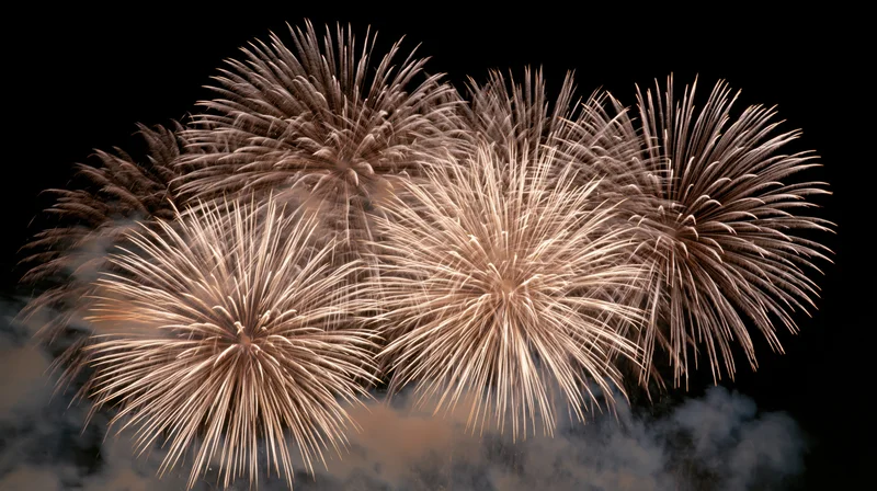Multiple fireworks bursting simultaneously against a black sky with smoke trails