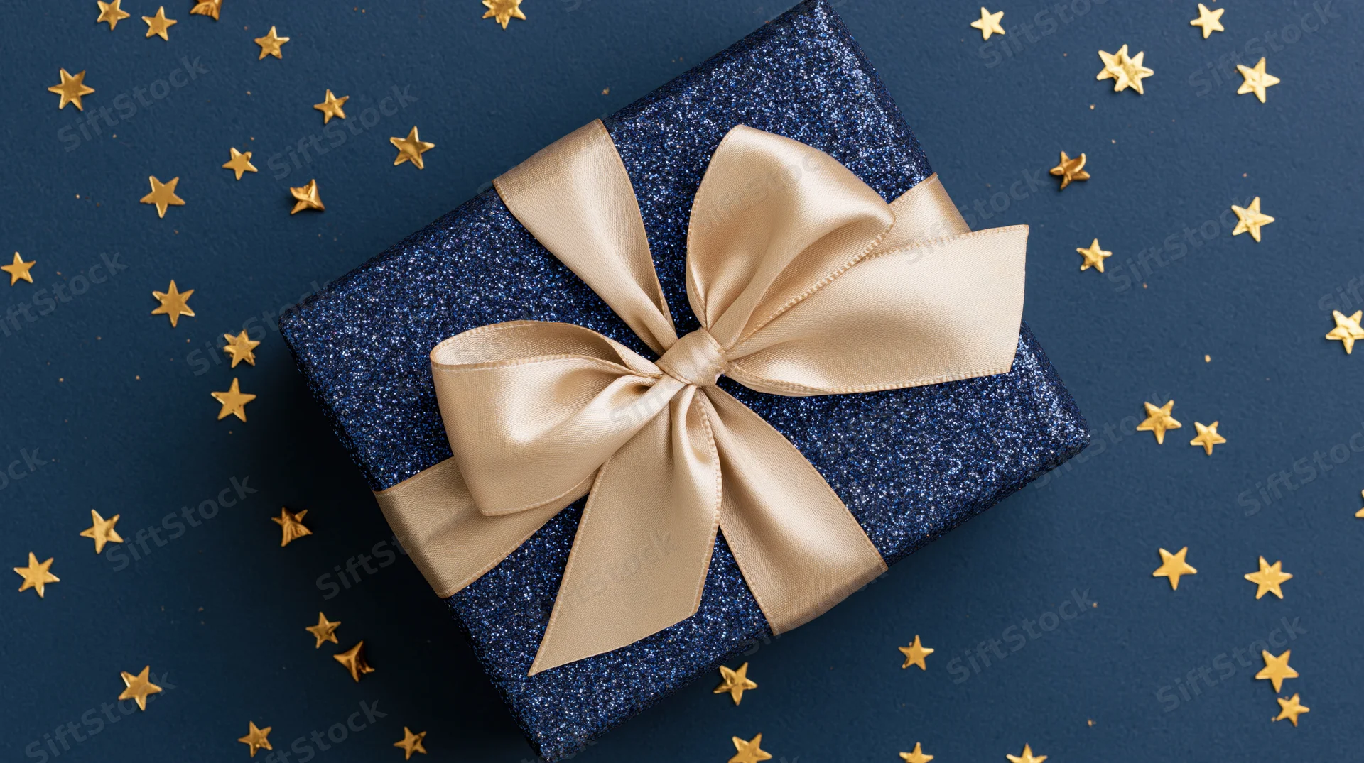 Navy blue glitter gift box with champagne satin bow on dark blue surface surrounded by gold stars