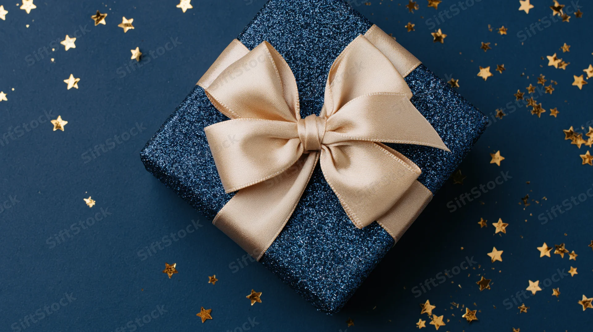 Navy blue glitter gift box with champagne satin bow on dark blue surface surrounded by golden star confetti