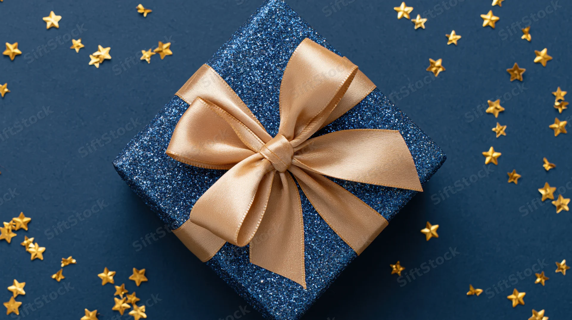 Navy blue glitter gift box with champagne satin bow surrounded by gold star confetti on dark blue surface
