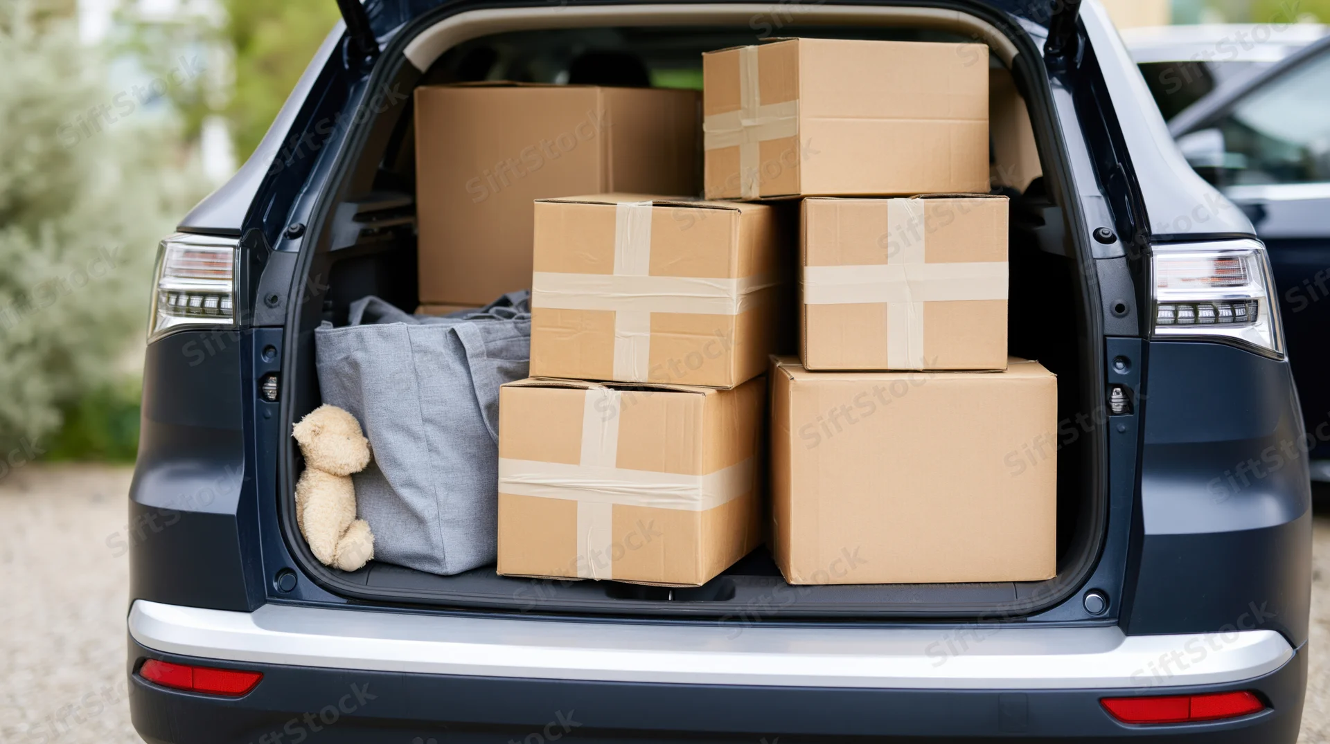 Open trunk of dark navy SUV filled with stacked cardboard boxes and a gray bag with a teddy bear inside, outdoors during daytime