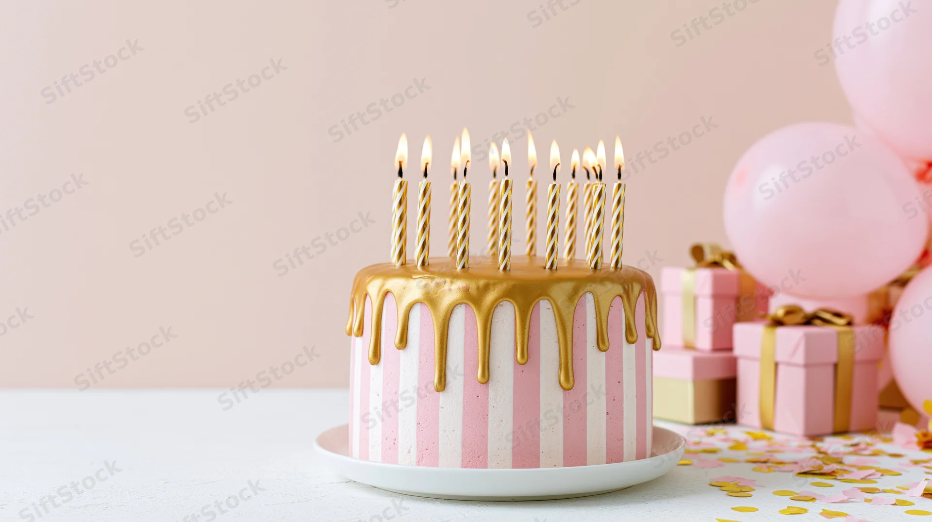 Pink and white striped birthday cake with gold drip frosting and lit candles on a table beside gifts and balloons