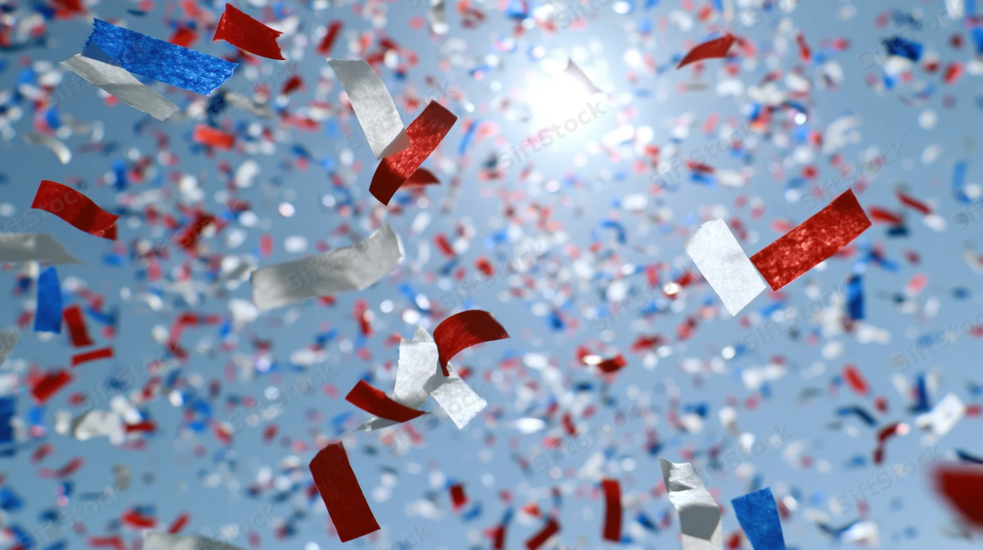 Red white and blue paper confetti falling against a bright blue sky with sun flare