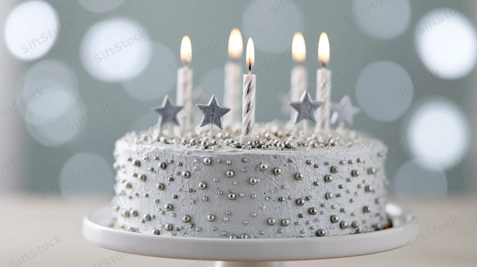 Silver-frosted birthday cake with lit candles and star decorations on a white cake stand