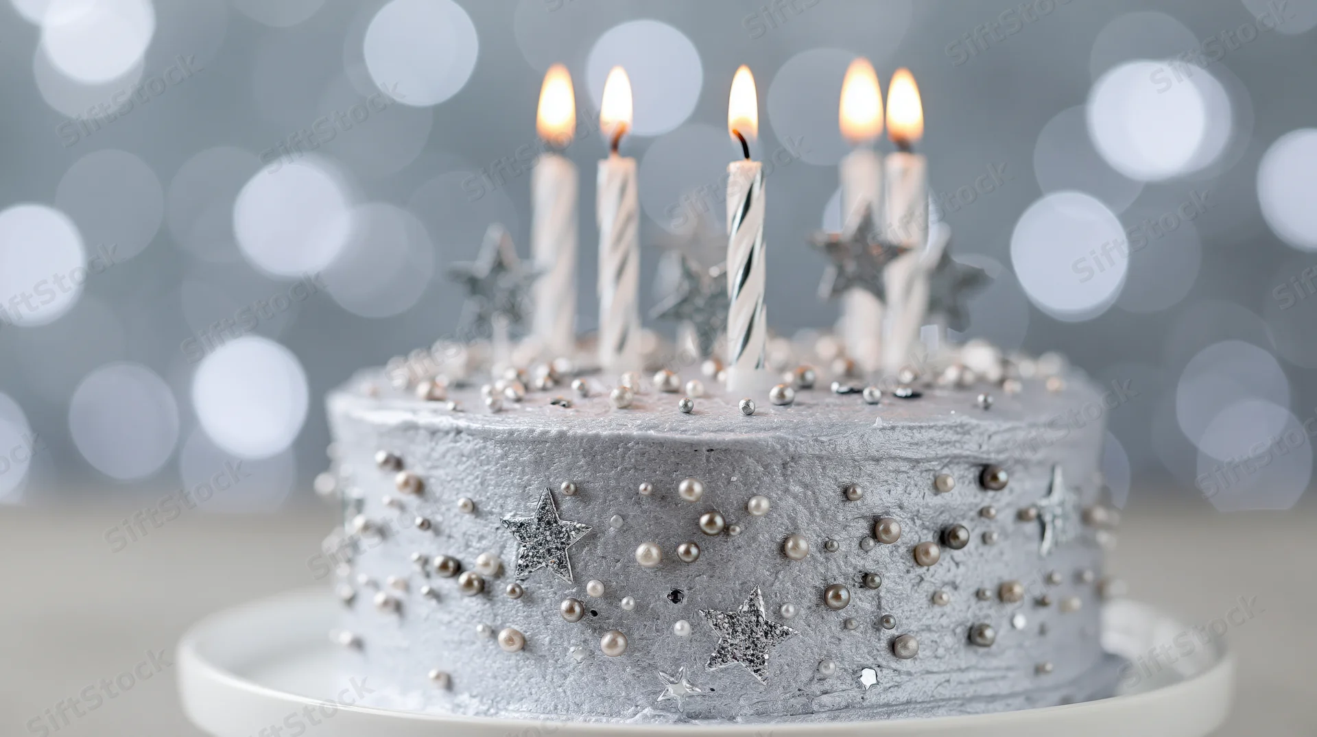 Silver-frosted birthday cake with lit candles and star decorations on a white stand