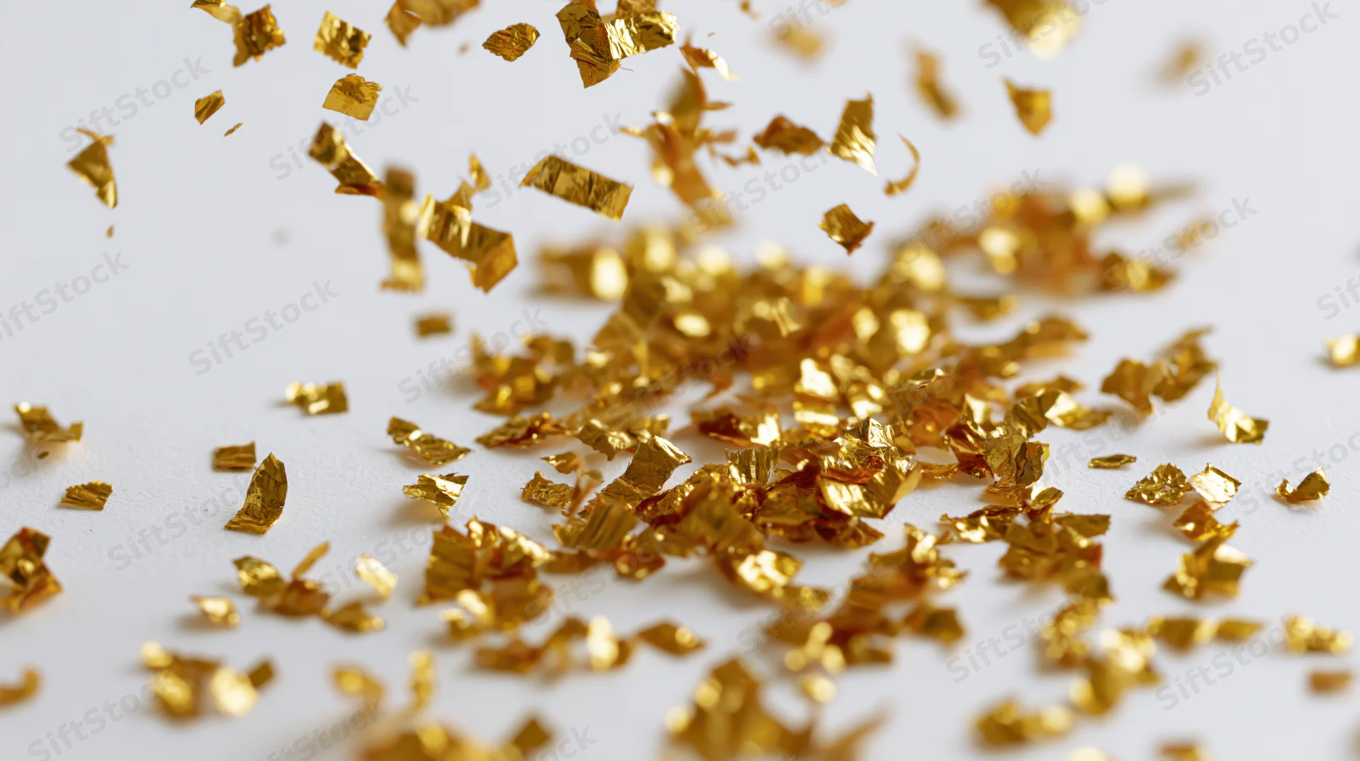 Gold metallic confetti pieces falling and tumbling midair on white surface