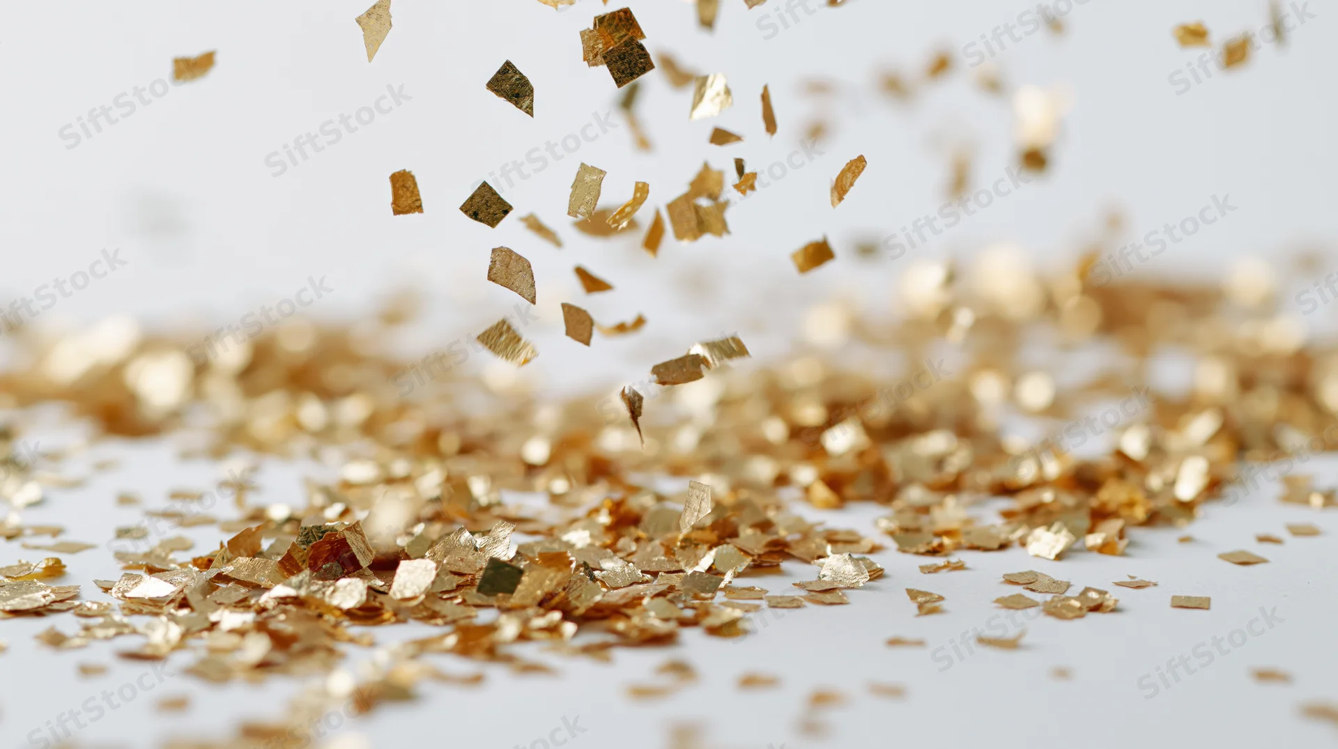 Gold metallic confetti pieces falling and tumbling mid-air over a white surface