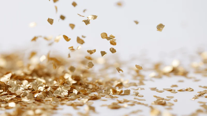 Gold metallic confetti pieces falling and tumbling midair over a white surface