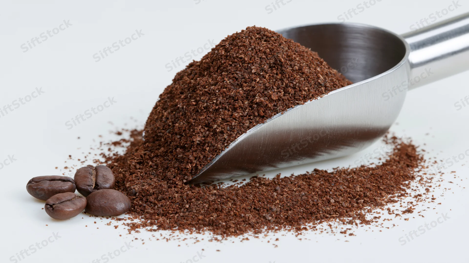 Stainless steel scoop heaped with ground coffee beside whole beans on white surface