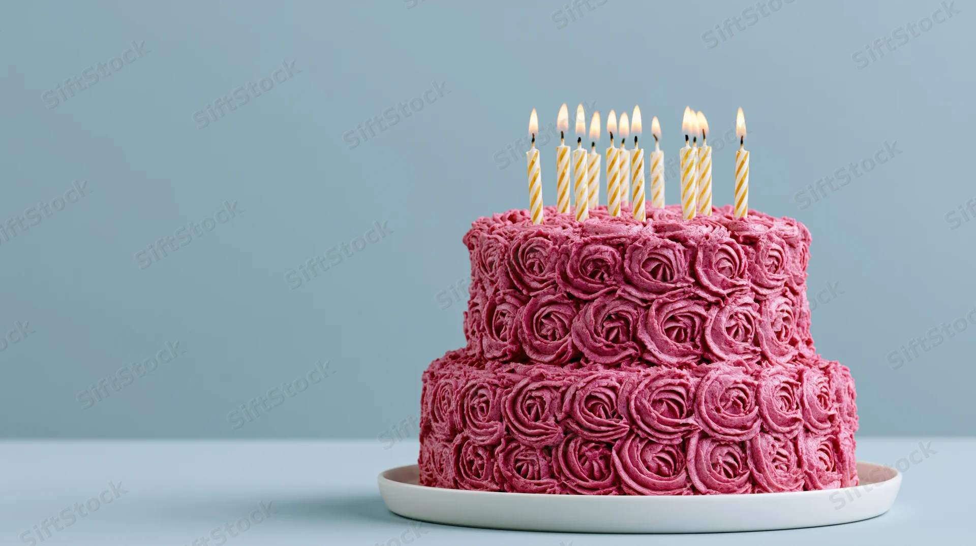 Two-tiered pink cake with rosette frosting and lit candles on a white plate against a blue background