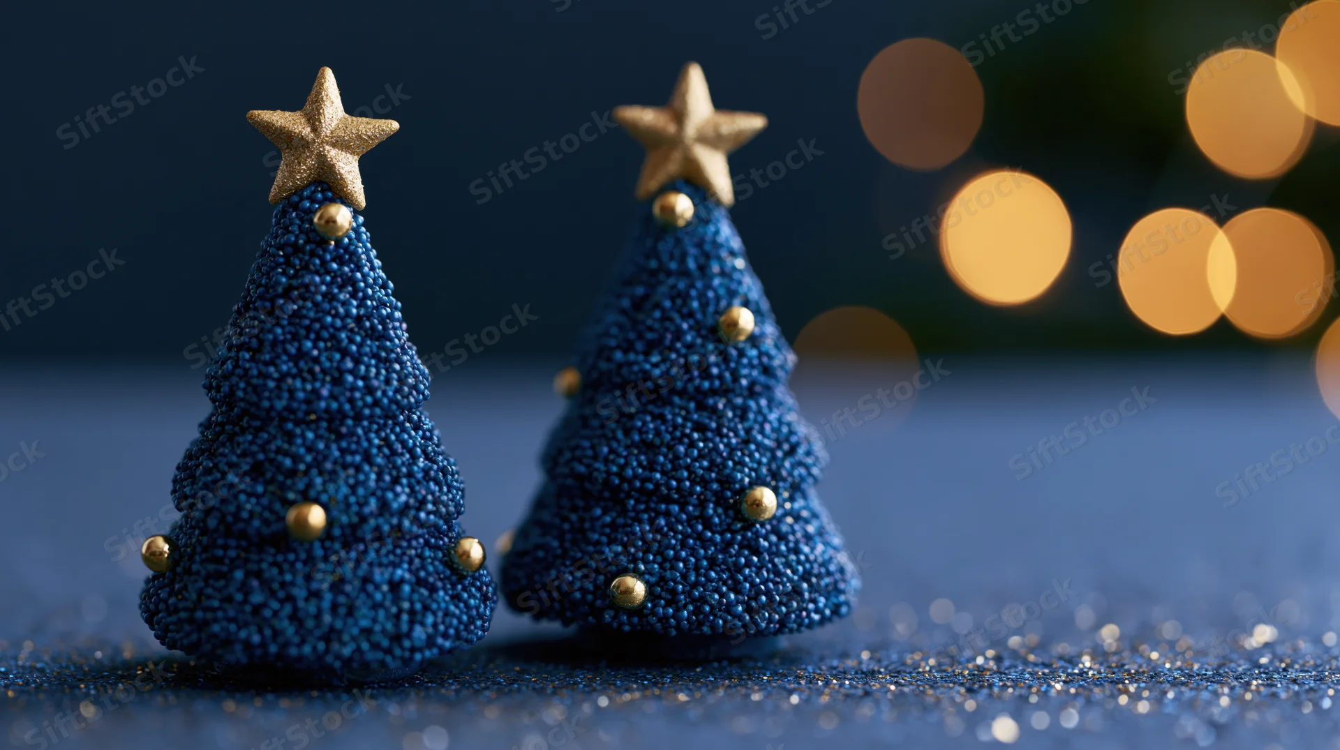 Two small blue beaded christmas trees with golden stars on a glittery surface against blurred warm lights