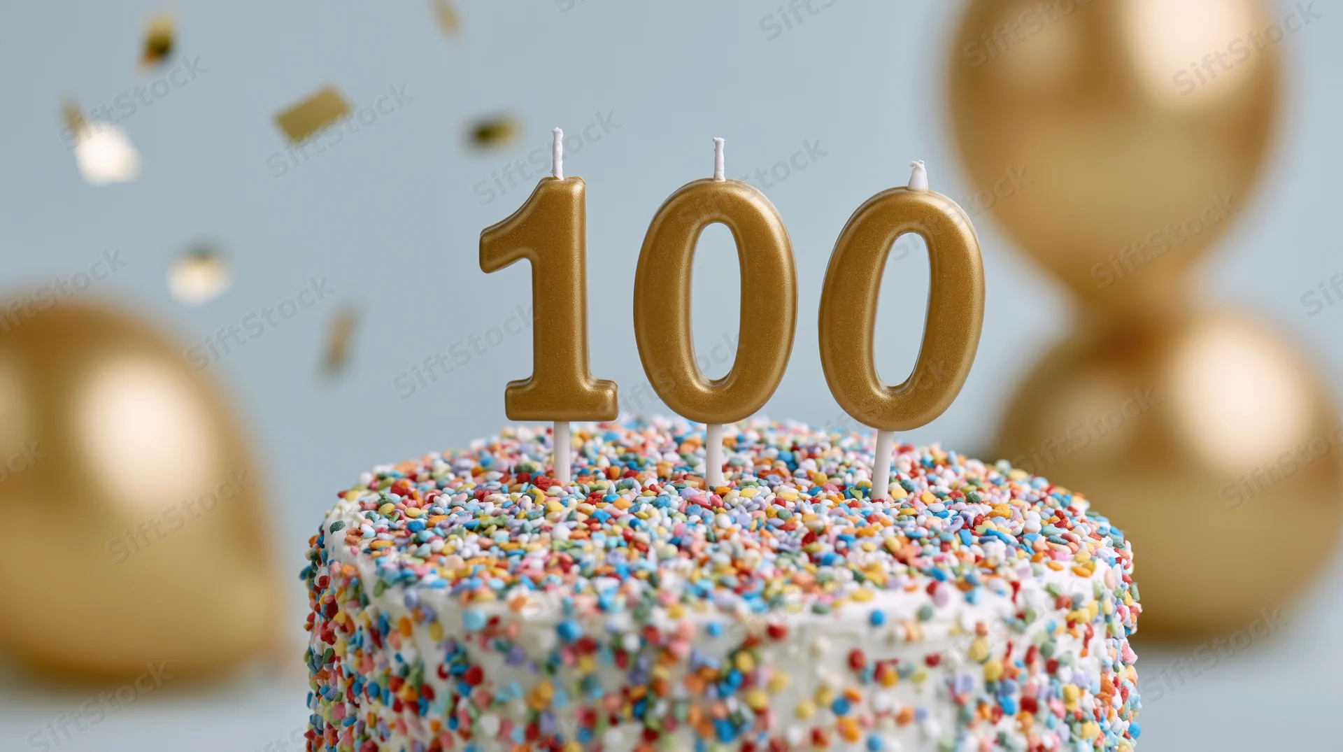 White cake covered in multicolored sprinkles with golden 100 candles and balloons in background