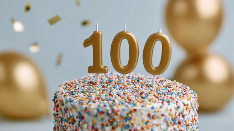 White cake covered in multicolored sprinkles with golden 100 candles and balloons in background