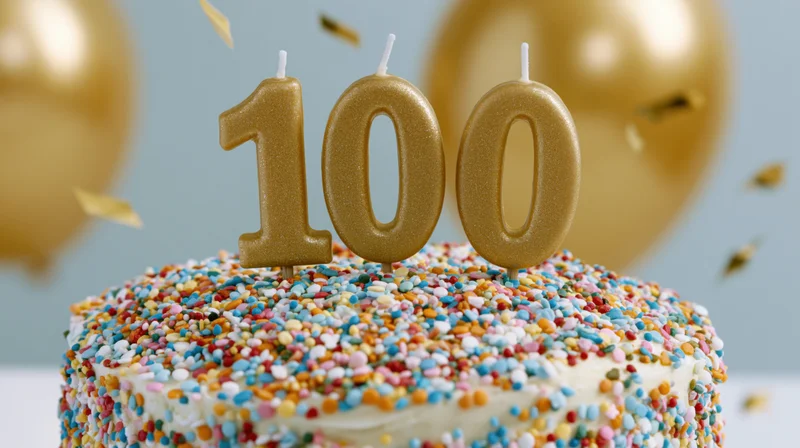 White cake covered in multicolored sprinkles with gold 100 candles and balloons in background