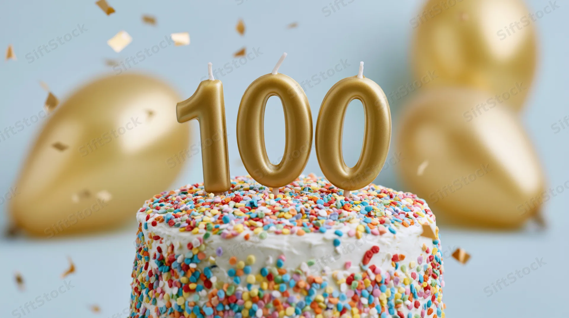 White cake with multicolored sprinkles and gold 100 candles surrounded by balloons and confetti