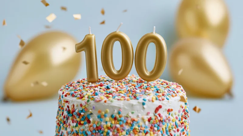 White cake with multicolored sprinkles and gold 100 candles surrounded by balloons and confetti