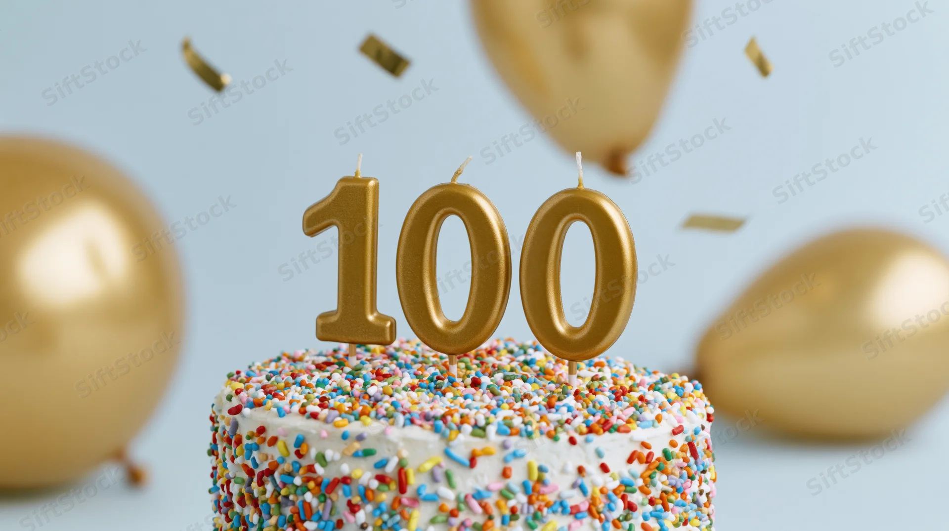White cake with gold 100 candles and sprinkles surrounded by balloons and confetti