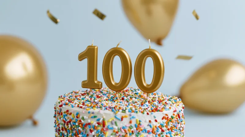 White cake with gold 100 candles and sprinkles surrounded by balloons and confetti