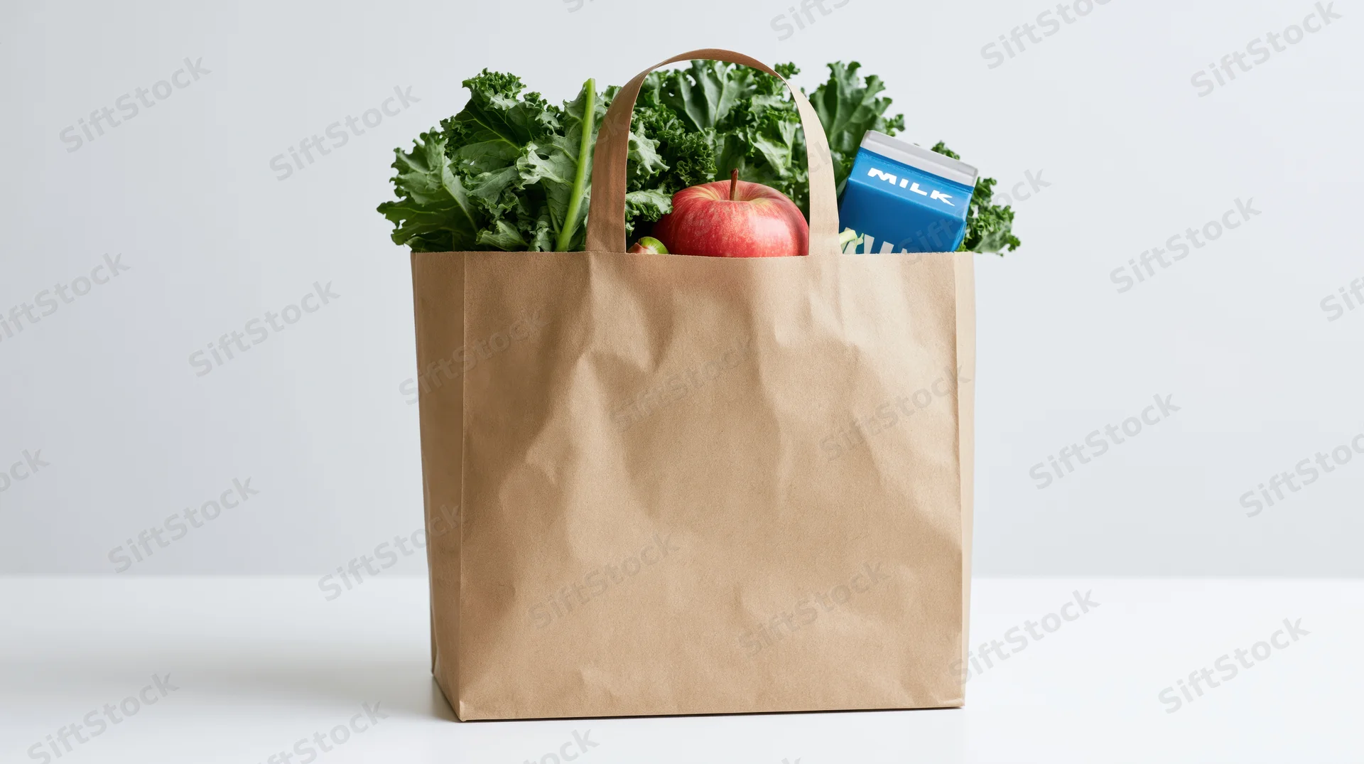 Brown paper grocery bag filled with fresh kale, apple, and milk carton on white surface