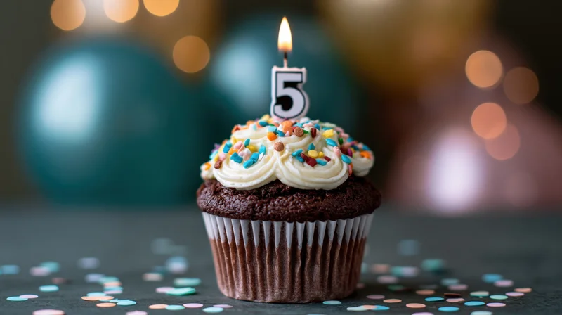 Chocolate cupcake with white frosting and number five candle surrounded by confetti on dark surface