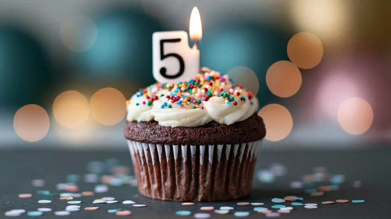 Chocolate cupcake with white frosting and number 5 candle on dark surface with scattered confetti and bokeh lights