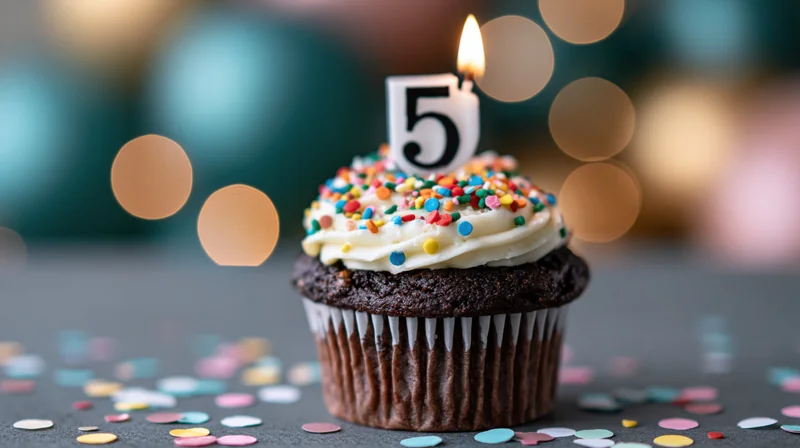 Chocolate cupcake with white frosting and number five candle surrounded by confetti on dark surface