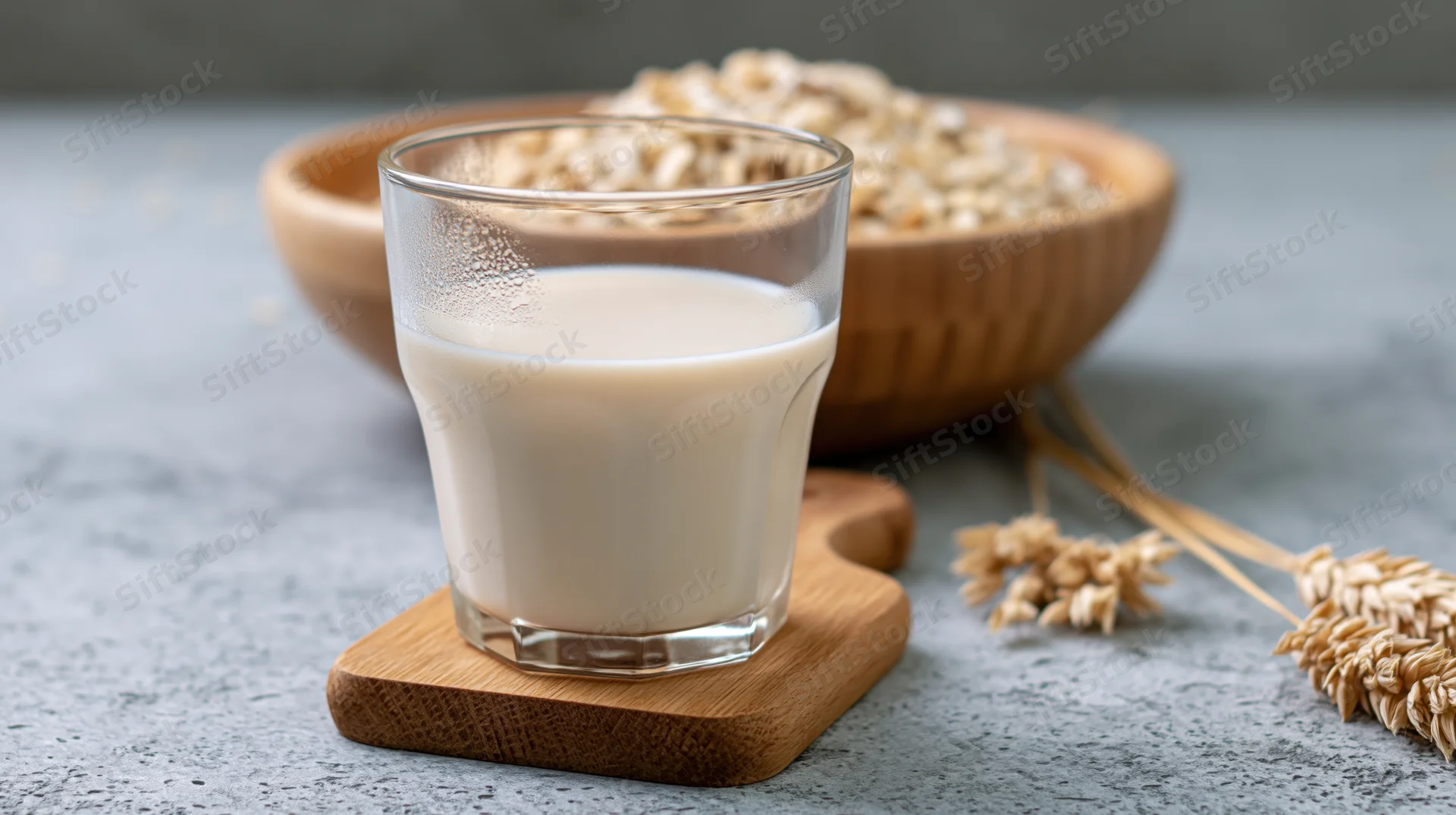 Glass of oat milk on wooden cutting board with oats in bowl and stalks nearby on gray surface