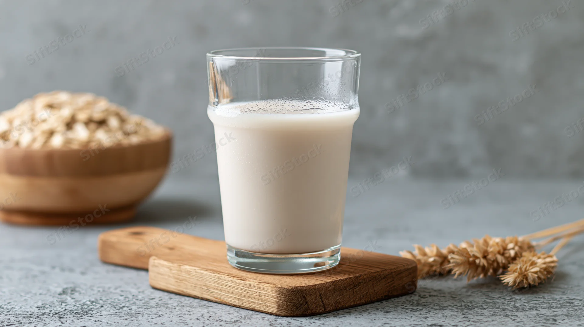 Glass of oat milk on wooden cutting board with oats and wheat stalks on gray surface