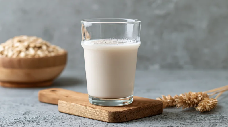 Glass of oat milk on wooden cutting board with oats and wheat stalks on gray surface