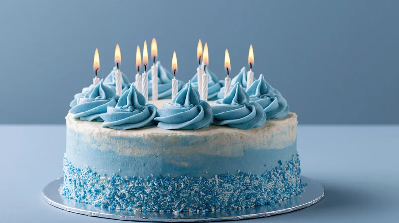 Blue and white frosted birthday cake with lit candles on a silver stand against a blue background