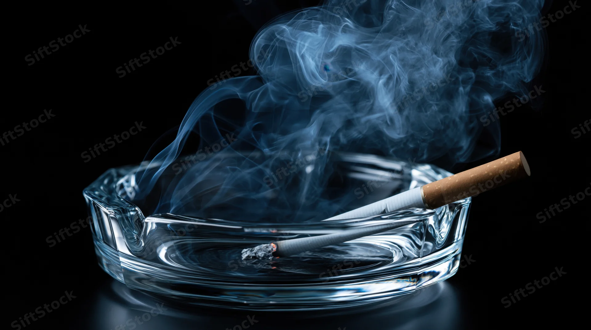 Lit cigarette resting in a clear glass ashtray with curling smoke against a black background