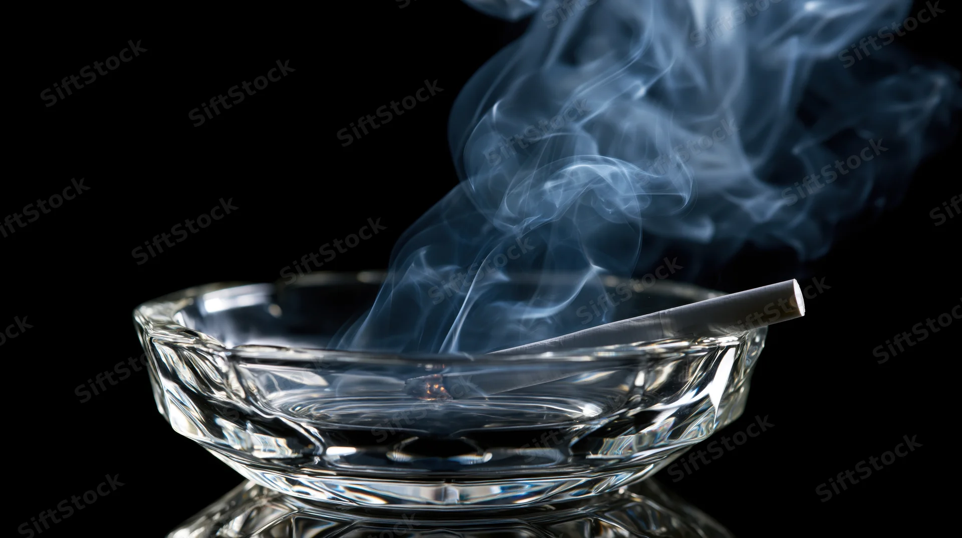 Lit cigarette in a clear glass ashtray with curling smoke against black background