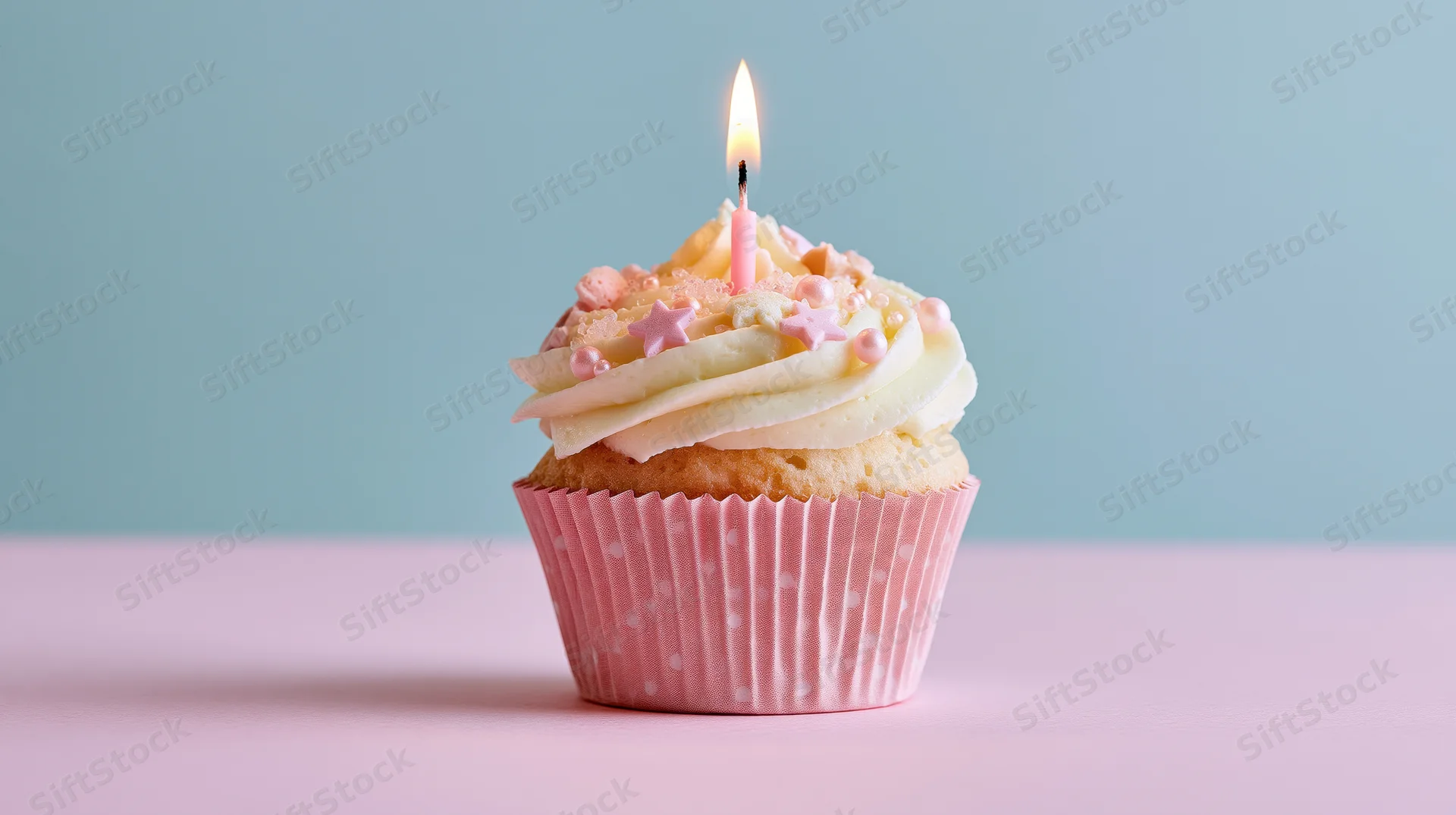 Single birthday cupcake with lit pink candle and white frosting on pink surface against blue background