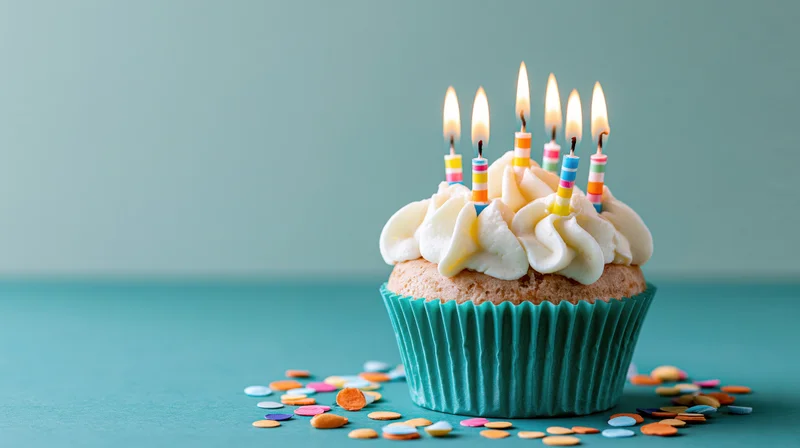 Birthday cupcake with lit candles and white frosting on teal surface surrounded by confetti