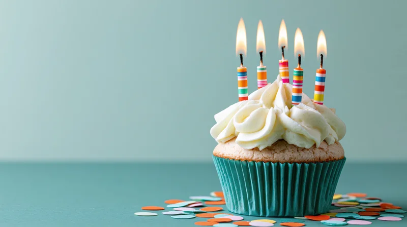 A birthday cupcake with white frosting and lit candles on a teal surface surrounded by confetti