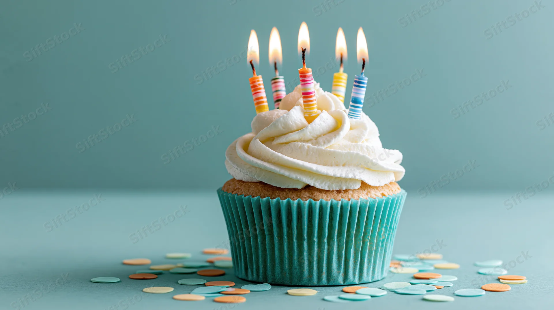 Birthday cupcake with lit candles and white frosting on teal surface surrounded by confetti