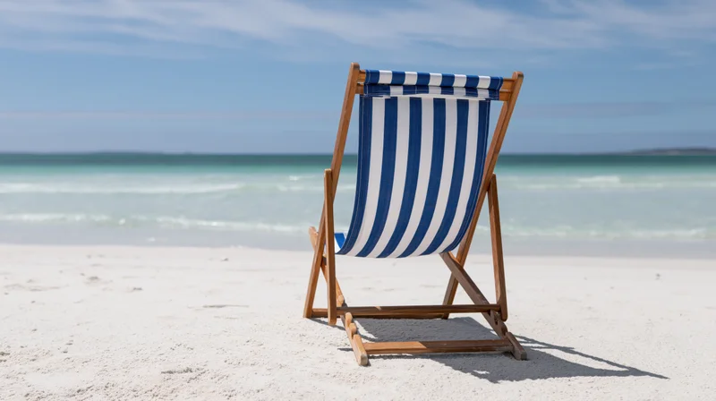 Blue and white striped wooden folding beach chair on sandy shore facing ocean under clear sky