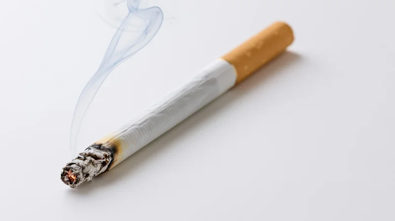 Lit cigarette burning diagonally on white surface with rising smoke