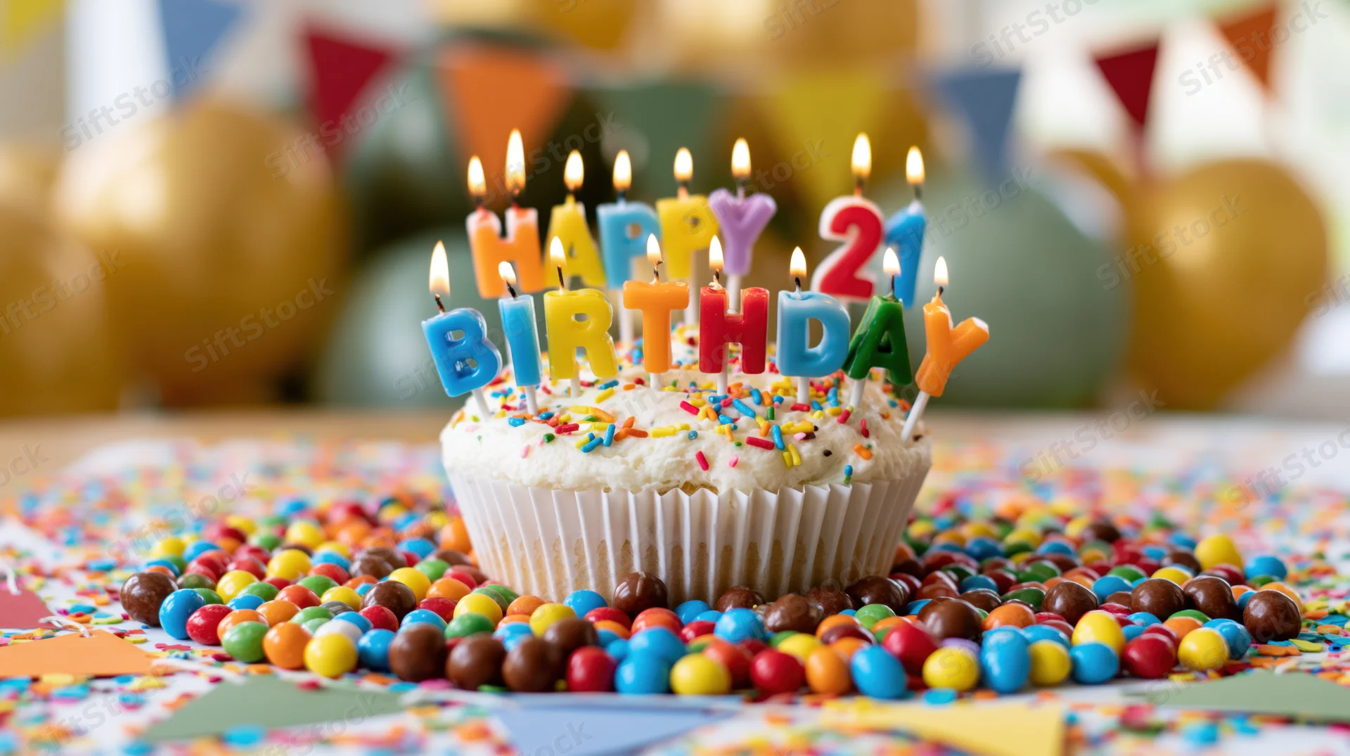 White-frosted cupcake with lit candles spelling happy birthday 21 surrounded by colorful candies and confetti on a party table