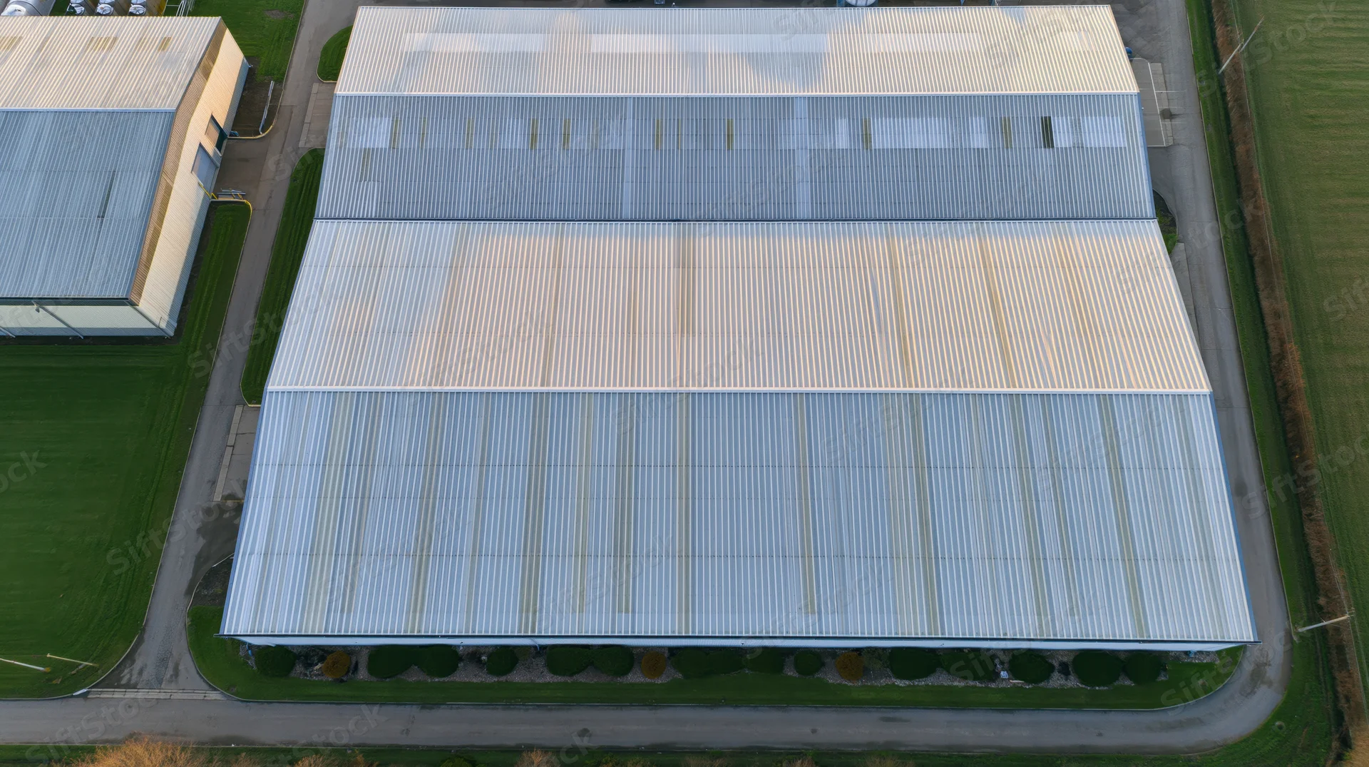 Aerial view of large industrial warehouse building with corrugated metal roof surrounded by grass and pavement