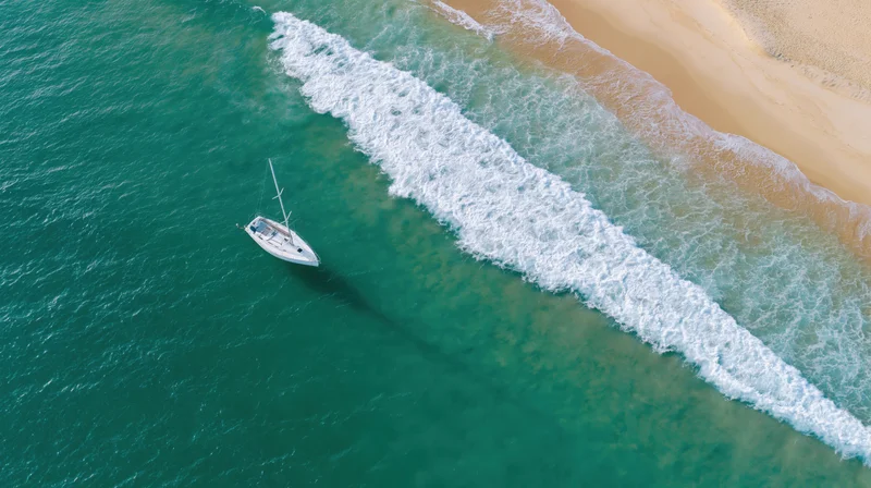 Aerial view of a white sailboat floating near a sandy beach with turquoise water and breaking waves