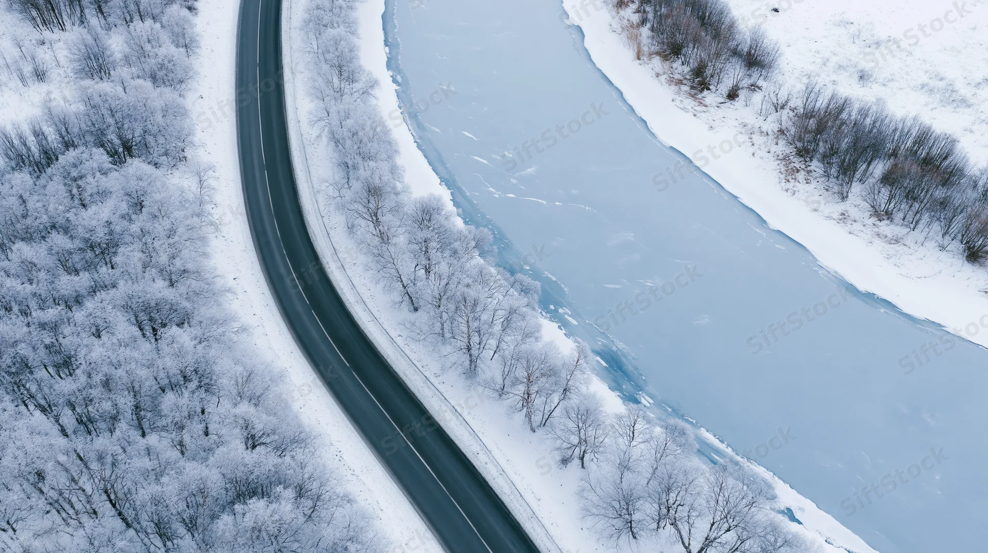 Winding road curving alongside frozen river through snow-covered forest from aerial view