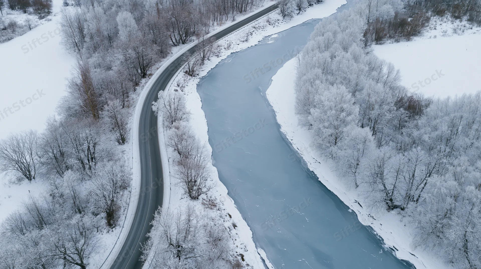 Aerial view of a winding road curving alongside a frozen river in a snow-covered forest landscape