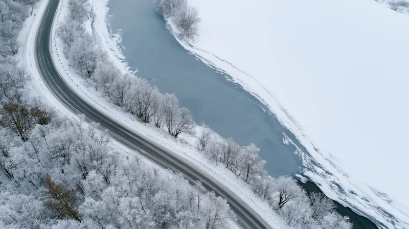Aerial view of a winding road curving alongside a frozen river through a snow-covered forest