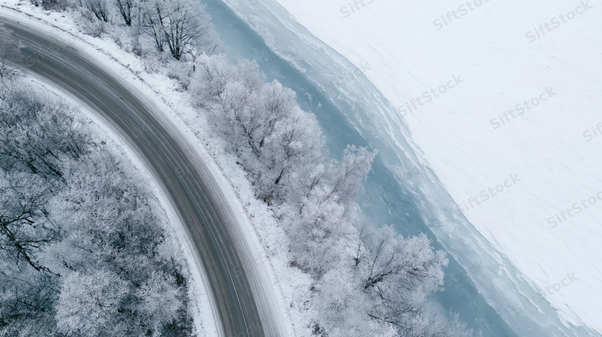 Aerial view of a winding road curving alongside a frozen river through snow-covered trees