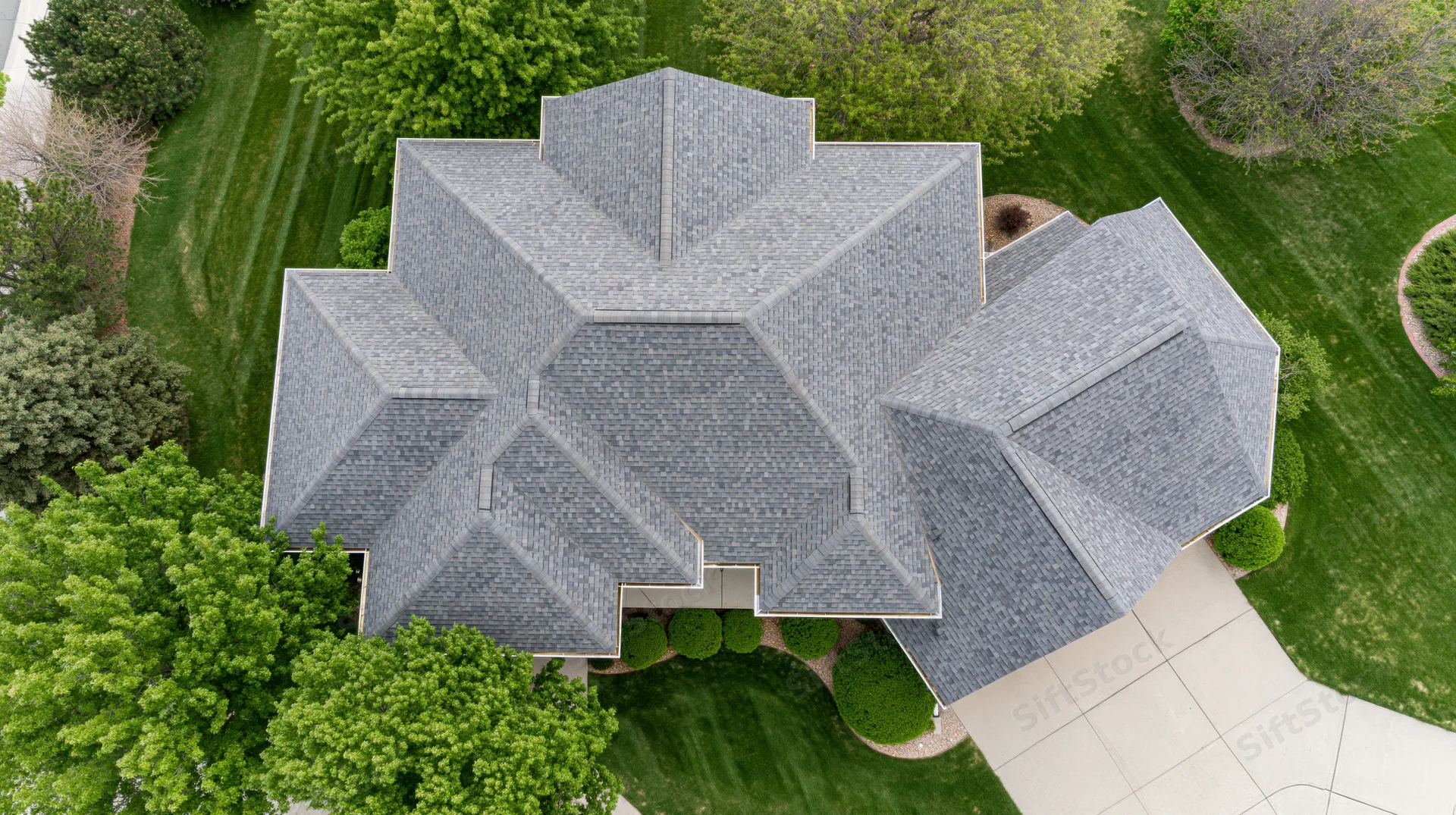 Aerial top-down view of a large suburban house with gray shingle roof surrounded by green lawn and trees