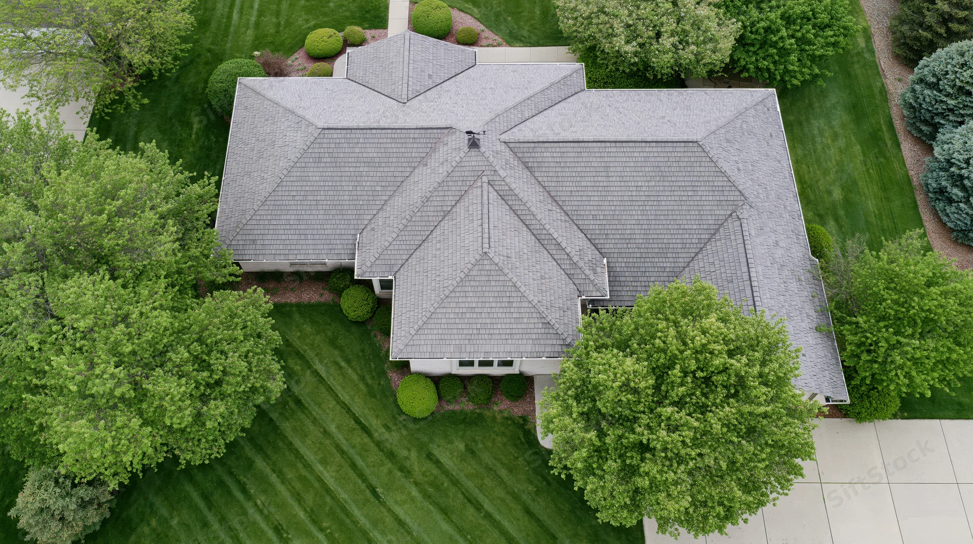 Aerial top-down view of a large suburban house with gray shingle roof surrounded by green lawn and trees