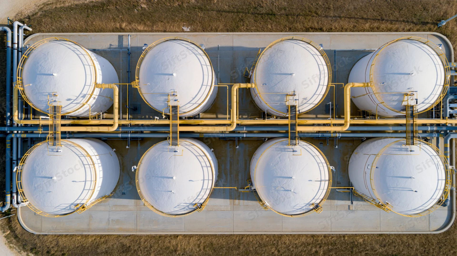Aerial top-down view of eight white cylindrical storage tanks arranged in two rows on a concrete platform surrounded by dry grass field