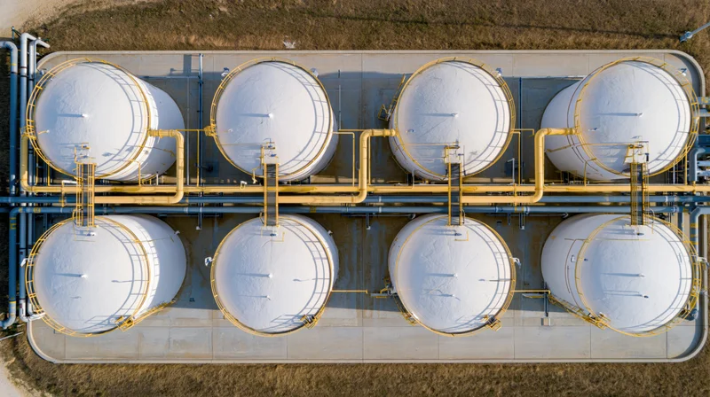 Aerial top-down view of eight white cylindrical storage tanks arranged in two rows on a concrete platform surrounded by dry grass field
